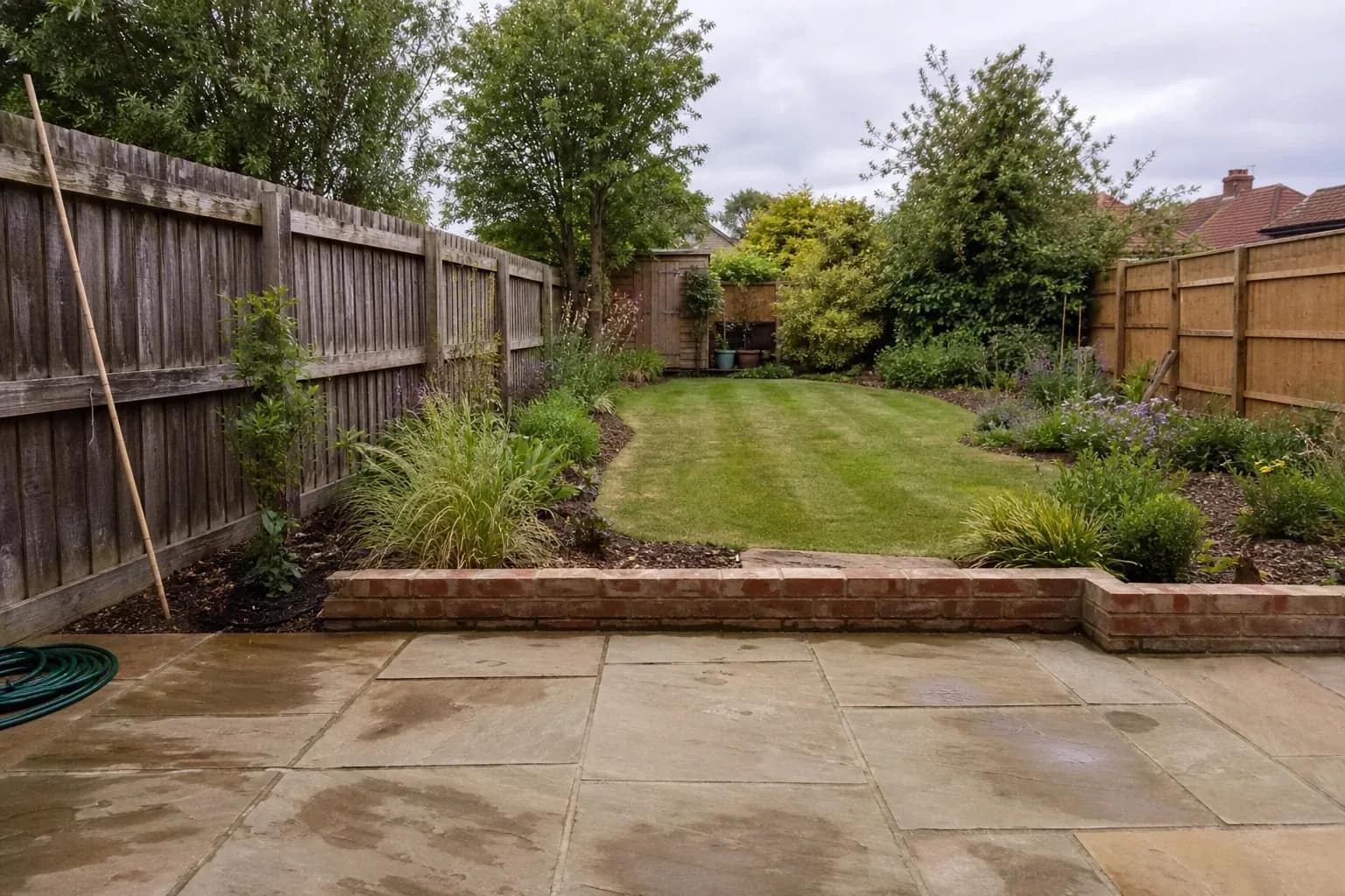 A modest suburban back garden with sandstone paving, part-grown borders and a weathered fence in summer light