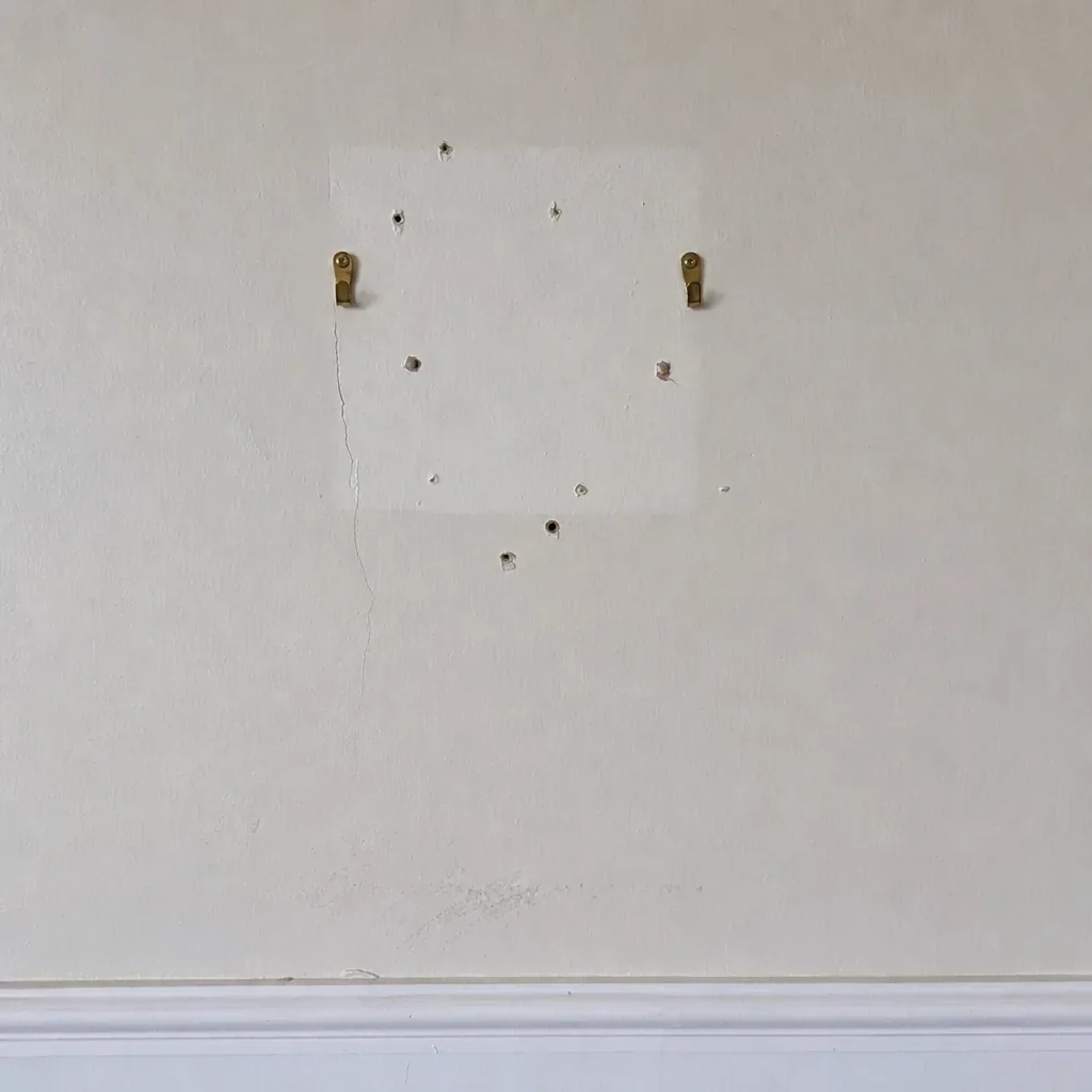 Close-up of brass picture hooks, nail holes and a cleaner rectangle of paint on an interior wall